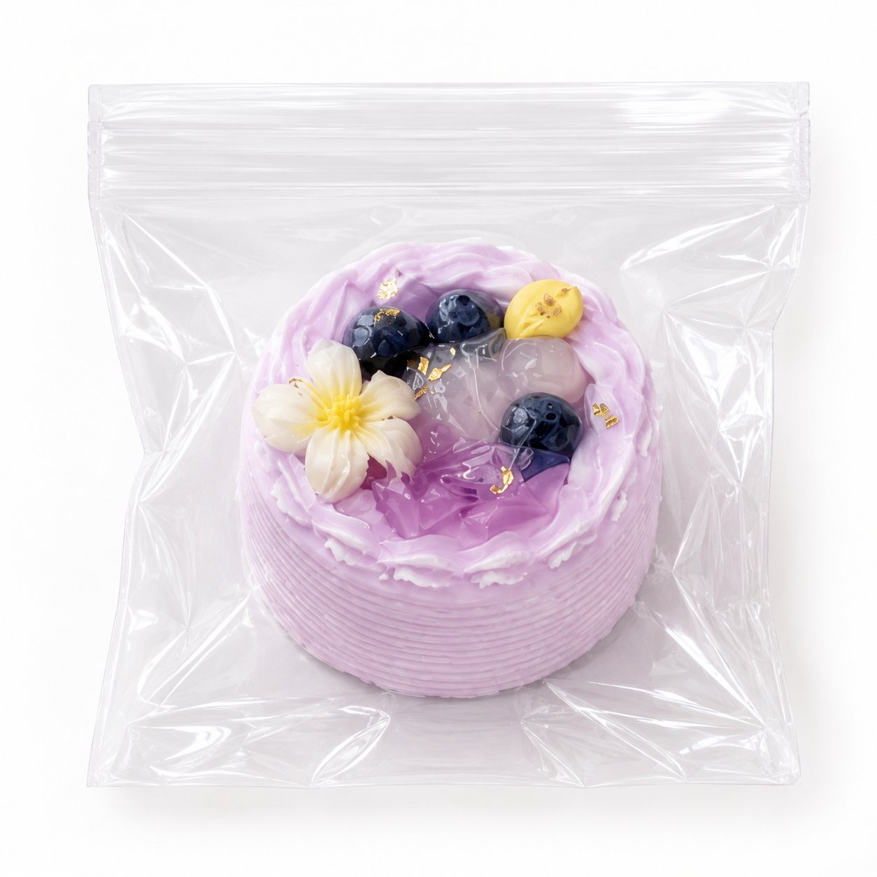 Purple blueberry and floral decorated mini cake squishy stress relief toy wrapped in clear plastic on a white surface.