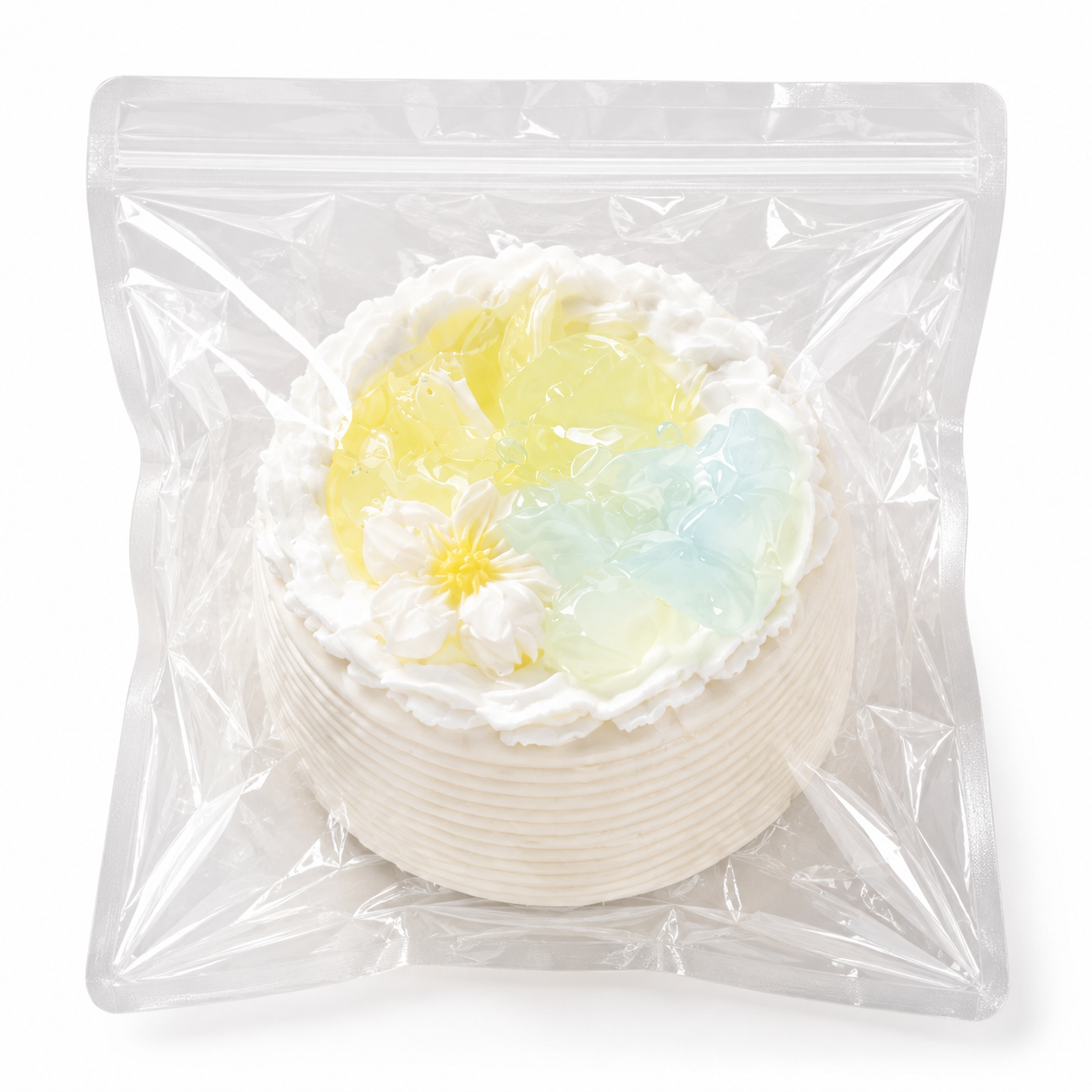 White mini cake squishy decorated with a daisy and pastel blue and yellow jelly pieces in a clear plastic bag.