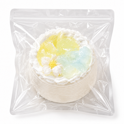 White mini cake squishy decorated with a daisy and pastel blue and yellow jelly pieces in a clear plastic bag.