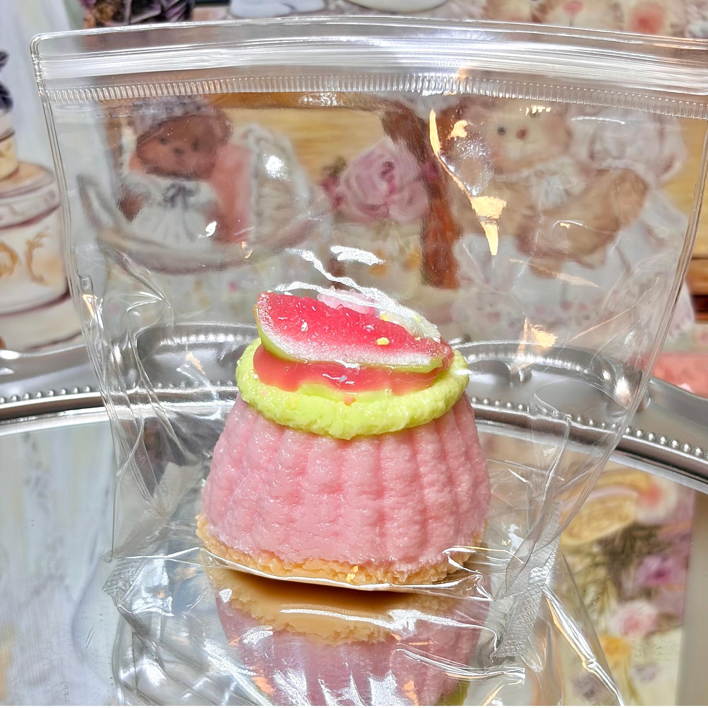 Pink cake-shaped squishy toy with watermelon topping in clear ziplock packaging on a silver tray.