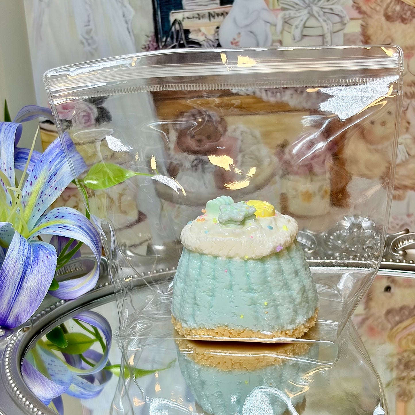 Blue pastel squishy dessert toy with star decorations packaged in a clear bag next to a blue lily.