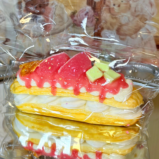 Close-up of a realistic yellow eclair squishy with red fruit and cream toppings in a clear plastic bag.