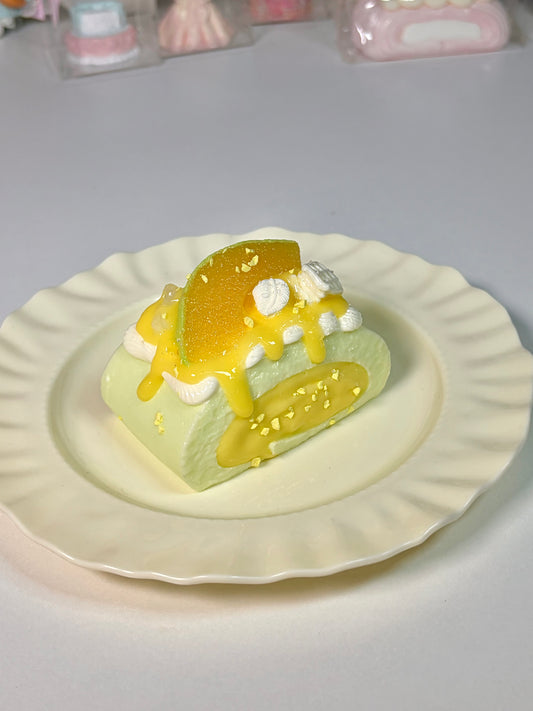 Green-base Swiss roll squishy with yellow mango slice topping, white cream, and glossy syrup on a wavy white plate over a plain white desk