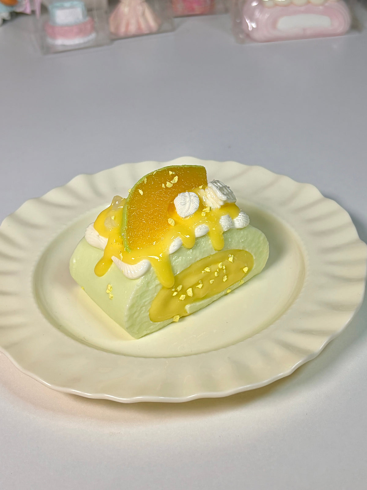 Green-base Swiss roll squishy with yellow mango slice topping, white cream, and glossy syrup on a wavy white plate over a plain white desk