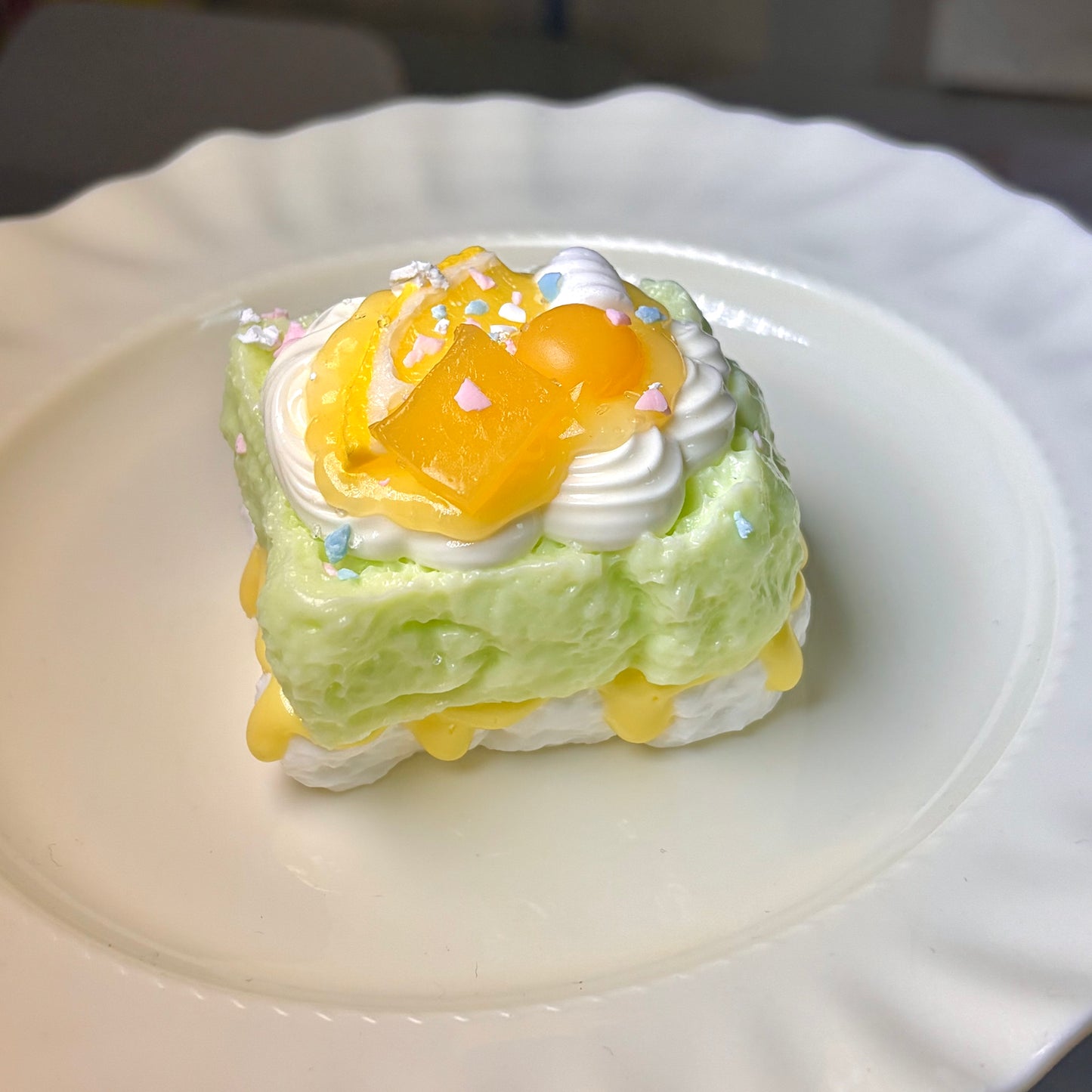 Green cube squishy with wrinkled texture, yellow syrup filling, white cream, and colorful sprinkle bits on a wavy white plate